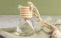 A small glass bottle with a wooden screw cap, attached to a decorative rope, likely used as a car fragrance diffuser. There's a fabric pouch in the background.